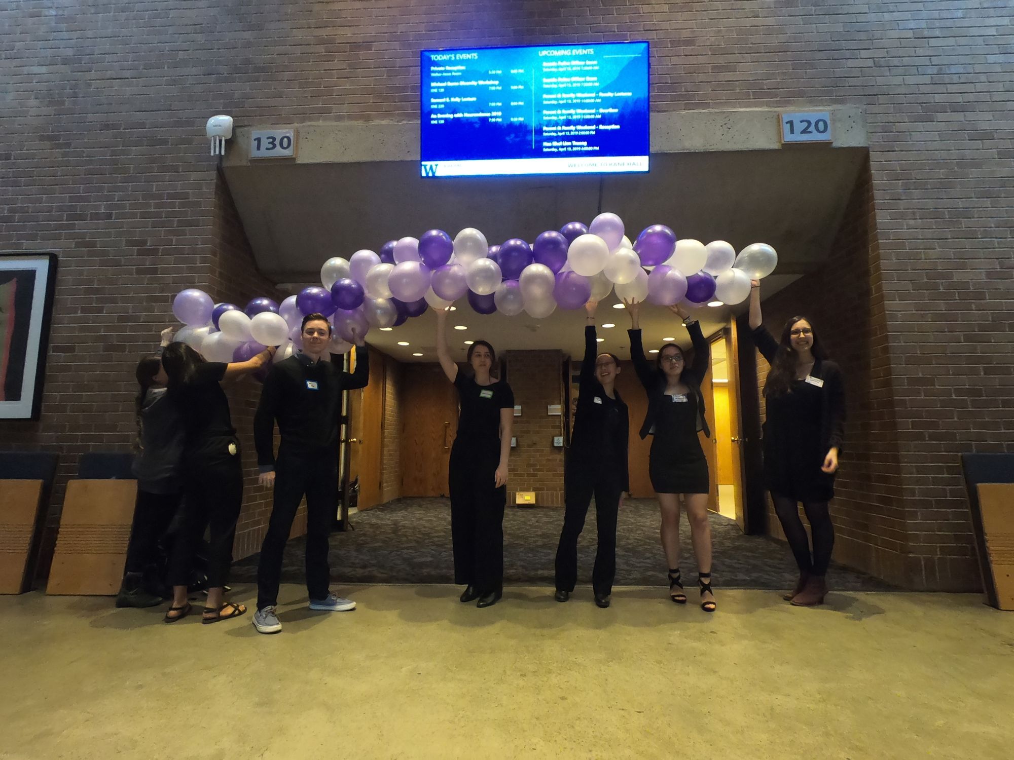 Members of the EWN planning committee, all dressed in black, hold a horizontally stretched purple and white balloon arch in Kane Hall's lobby