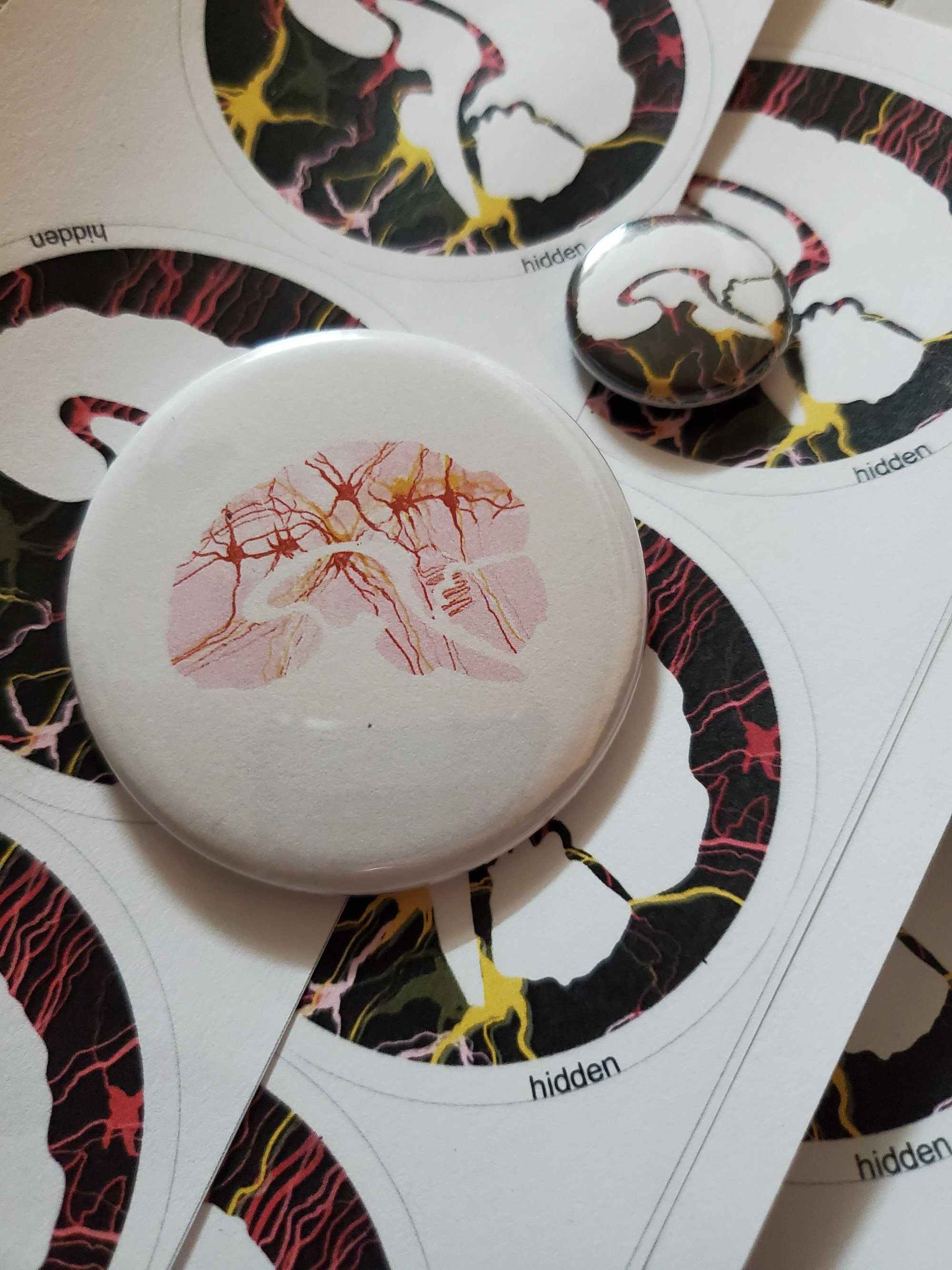 A circular white button pin with a pink brain on it lays on top of pages of circular EWN stickers with white brains on black backgrounds.